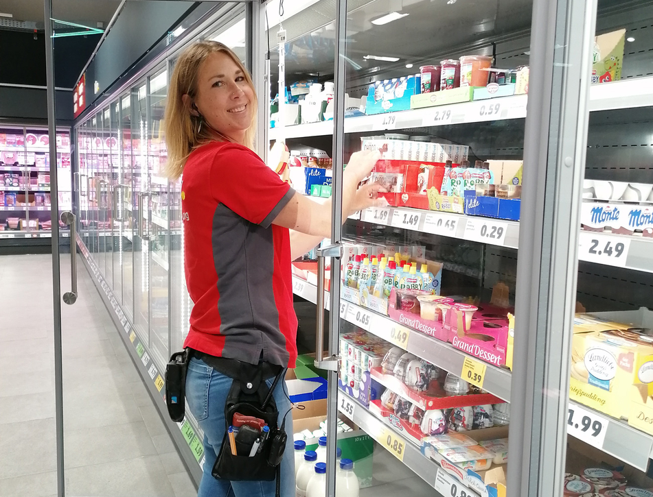 Frederike Elison befüllt lächelnd ein Kühlregal in einem PENNY Markt und blickt in die Kamera