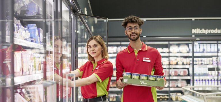 Eine PENNY Mitarbeiterin und ein Mitarbeiter stehen in einem Markt vor einem Kühlregel nebeneinander. Die linke Mitarbeiterin hebt etwas aus dem Kühlregal. Der rechte Mitarbeiter trägt eine Palette mit Lebensmitteln.