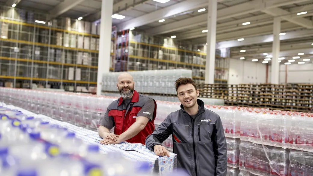 Zwei PENNY Mitarbeiter lehnen in Logistikkleidung an Produktpaletten in einem Logistik Standort