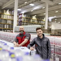 Zwei PENNY Mitarbeiter lehnen in Logistikkleidung an Produktpaletten in einem Logistik Standort