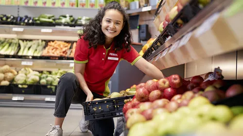 Eine PENNY Mitarbeiterin in einem Markt hebt eine Obst und Gemüse Kiste in ein Regal